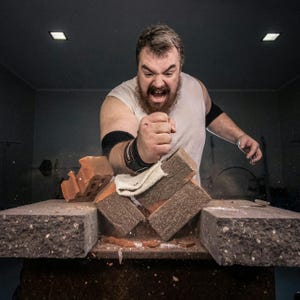 May include: A person in a white tank top breaks several red bricks with a fist. The bricks are on a dark gray surface, and the person is wearing black wrist and elbow supports. A white cloth is on the bricks.