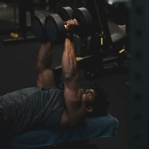 May include: A person lying on a bench in a gym, lifting two black dumbbells. The person is wearing a gray tank top. The dumbbells appear to be approximately 12 inches long.