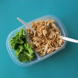 May include: A clear plastic divided container filled with a meal of shredded chicken and rice, and broccoli florets. Two white plastic forks are placed in the container. The container sits on a textured teal surface.