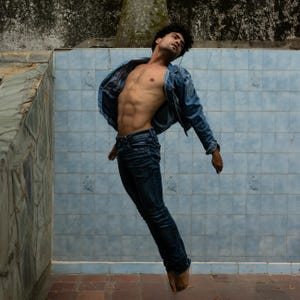 Puede incluir: Una persona en el aire, vistiendo una chaqueta vaquera y vaqueros azul oscuro. La persona está sin camisa, con un físico musculoso. El fondo presenta una pared de azulejos azules y una estructura de piedra.