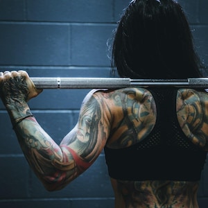 May include: A person with detailed tattoos on their arms and back, holding a barbell across their shoulders. They are wearing a black sports bra. The background is a dark blue brick wall, suggesting a gym or workout setting.
