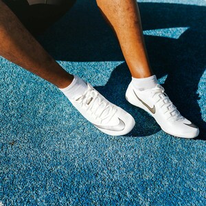 May include: A pair of white athletic cleats featuring a metallic silver Nike swoosh logo on the side, white laces, and white ankle socks. The footwear has a streamlined, low-profile design with a textured surface, visible on a bright blue track.