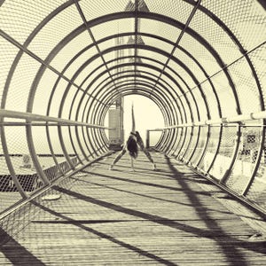 Op de afbeelding: Een sepia-getinte afbeelding van een persoon die een yogahouding uitvoert in een metalen tunnel. De tunnel heeft een cirkelvormig ontwerp met een houten vloer en metalen relingen. De persoon draagt een legging met patroon en een top.