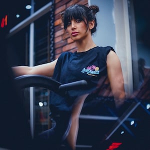 May include: A black sleeveless t-shirt with the text "Iron Maniels" in a stylized script, featuring a pink pineapple and palm trees. The shirt is worn by a person in a gym setting, with a brick wall visible in the background.