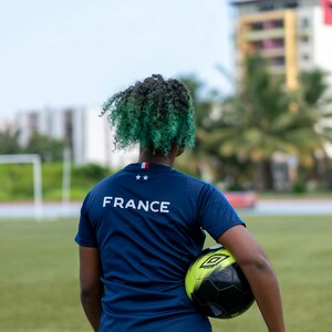May include: A person wearing a navy blue t-shirt with the word "FRANCE" printed on the back, holding a black and neon green soccer ball. The person has green and black curly hair. The image is taken on a green field.