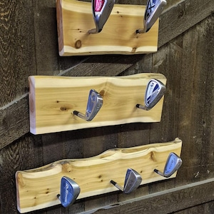 May include: Three horizontal live-edge wooden wall racks mounted vertically. Each rack features two or three silver metal golf club heads attached as hooks. The natural light-colored wood displays visible grain patterns and knots, contrasting with the dark wood background.