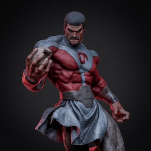 Può includere: Un'action figure maschile muscolosa con baffi scuri e capelli corti, che indossa una tuta rossa e grigia con un emblema circolare sul petto. La figura presenta una cintura grigia e un indumento grigio fluente simile a una gonna, tenendo una pietra.