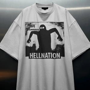 Può includere: Una t-shirt a maniche corte grigio chiaro con una stampa grafica nera di una figura ombrosa con due occhi bianchi. Il testo "YOUR CHAOS DAYS ARE NUMBERED" appare in alto a sinistra e la parola "HELLNATION" è stampata in grassetto bianco sotto.