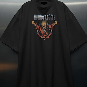 Puede incluir: Camiseta negra con la palabra "THUNDERDOME" sobre un gráfico de un personaje con una túnica roja con los brazos levantados. El gráfico está dentro de un círculo azul y rojo. La camiseta tiene mangas cortas y cuello redondo.