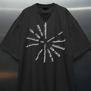 Może przedstawiać: Czarny t-shirt z krótkim rękawem z białym nadrukiem graficznym na klatce piersiowej. Wzór składa się z wielu krótkich, teksturowanych białych linii ułożonych w okrągły, promienisty kształt, rozchodzący się z centralnego punktu tkaniny.