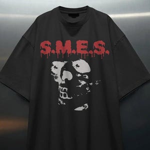 May include: A black short-sleeved t-shirt featuring a red graphic print of the text S.M.E.S. in a dripping font style above a white, high-contrast, stylized portrait graphic of a face with distinct features centered on the chest area.