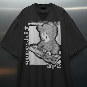 Puede incluir: Una camiseta negra de manga corta con un estampado gráfico rectangular blanco en el pecho. El gráfico muestra una figura parecida a un oso tocando un teclado, con el texto "goreshit" impreso verticalmente a la izquierda y "ap2" abajo a la derecha.