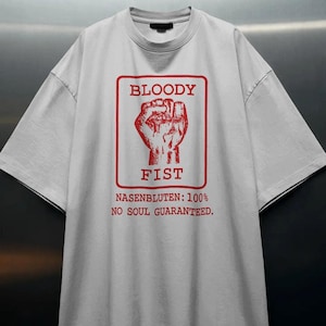 May include: A light grey short-sleeve t-shirt featuring a red rectangular graphic with a fist illustration. The text reads BLOODY FIST, NASENBLUTEN: 100%, and NO SOUL GUARANTEED. The shirt has a crew neckline and a relaxed fit with wide sleeves.