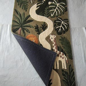 May include: A rectangular tufted rug with an olive green background featuring a jungle theme. The design includes a white winding path, a giraffe, and a partial lion face surrounded by dark green palm and monstera leaves. One corner is folded back.