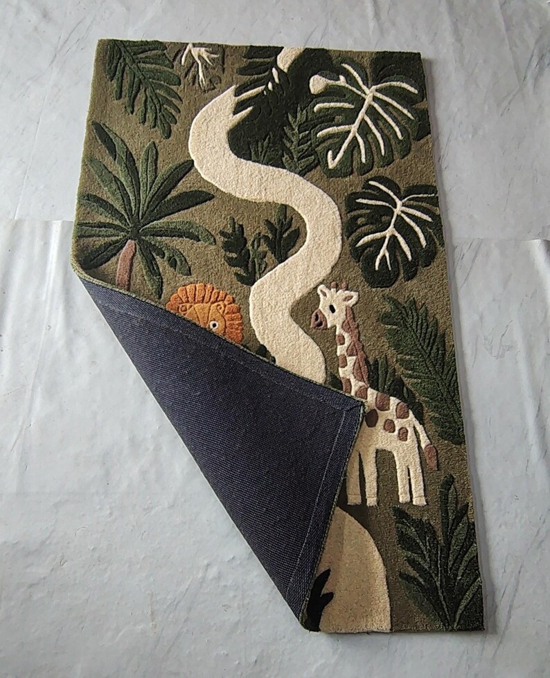 May include: A rectangular tufted rug with an olive green background featuring a jungle theme. The design includes a white winding path, a giraffe, and a partial lion face surrounded by dark green palm and monstera leaves. One corner is folded back.