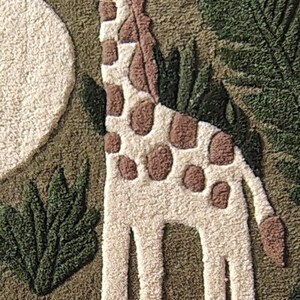 May include: A tufted textile wall hanging featuring a cream-colored giraffe with brown spots standing amidst dark green foliage and textured cream shapes. The design uses a punch needle technique to create a raised, tactile surface with distinct patterns.
