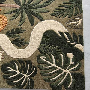 May include: A rectangular tufted rug featuring an olive green background with a large, winding cream-colored snake. The design includes dark green monstera leaves, palm fronds, and a textured orange circular element on the left side.
