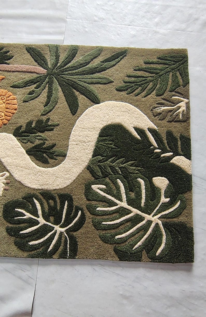 May include: A rectangular tufted rug featuring an olive green background with a large, winding cream-colored snake. The design includes dark green monstera leaves, palm fronds, and a textured orange circular element on the left side.