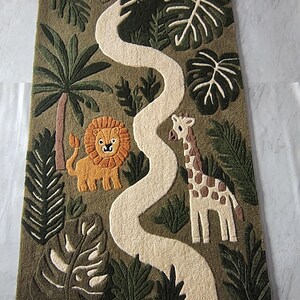 May include: A rectangular tufted rug featuring a jungle scene with a winding cream-colored path. The design includes a yellow lion with an orange mane, a cream and brown giraffe, and various green palm leaves and trees on an olive green background.