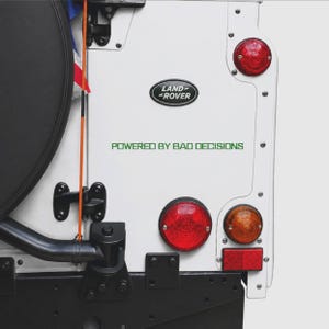 May include: Close-up of a white Land Rover vehicle's rear panel. Features the Land Rover logo, red and amber lights, and the text "POWERED BY BAD DECISIONS" in green.