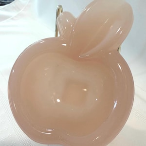 May include: A light pink glass apple-shaped dish with a stem and leaf design. The dish has a smooth, glossy surface and is likely used for decorative purposes or serving small items. The apple dish is set against a white background.