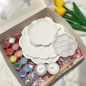 May include: A craft kit in an open box, featuring white ceramic plates, a paint palette, and small paint pots in various colours. Includes varnish, primer, and paintbrushes. The box is filled with red shredded paper. Yellow tulips are in the background.