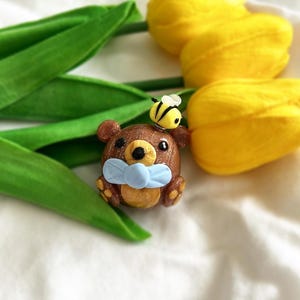 Puede incluir: Una figura artesanal de oso marrón con una pajarita azul claro y una abeja amarilla y negra en la cabeza. El oso está colocado cerca de tulipanes amarillos y hojas verdes sobre un fondo blanco.