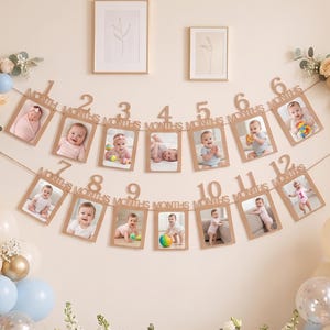 Puede incluir: Una guirnalda de fotos para hitos de bebé con doce marcos de madera numerados del 1 al 12 meses, colgados de una cuerda. La decoración incluye racimos de globos azul claro, crema y dorados, junto a un arreglo floral de rosas y follaje.