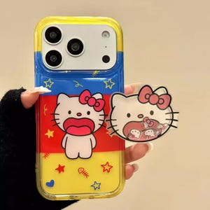 Puede incluir: Una funda de teléfono amarilla, azul y roja con un diseño de gato de dibujos animados. La funda tiene un borde transparente e incluye gráficos de estrellas y espinas de pescado. Un dije a juego con forma de gato se sostiene junto al teléfono.