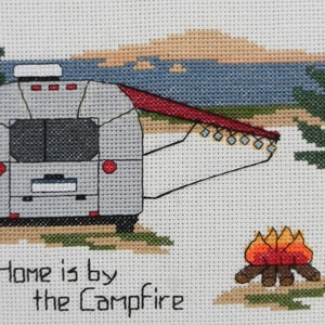 May include: Cross-stitch design featuring a silver camper with a red awning, a campfire, and the text "Home is by the Campfire." The scene includes trees, a lake, and a sunset in the background. The design is on a white background.