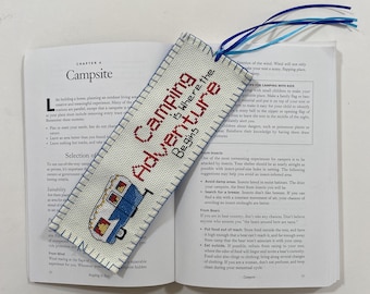 Op de afbeelding: Een witte bladwijzer met een blauw en rood ontwerp van een camper. De bladwijzer is gecross-stitched en heeft de tekst "Camping is Where the Adventure Begins".
