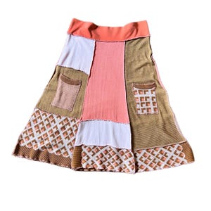 May include: A knee-length skirt featuring a patchwork design with panels of solid coral, light pink, and textured brown fabric. The skirt includes two patch pockets and a geometric diamond pattern in white, brown, and coral along the hemline.
