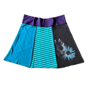 May include: A multi-paneled skirt featuring a wide purple waistband. The skirt consists of a solid teal panel, a central panel with teal and black horizontal stripes, and a black panel on the right side decorated with a blue floral graphic print.