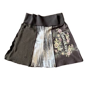 May include: A skirt featuring a wide dark brown waistband and three distinct panels. The left panel has white vertical pinstripes, the center panel displays a white and beige abstract brushstroke pattern, and the right panel shows floral graphics with the word CHOCOLATE.