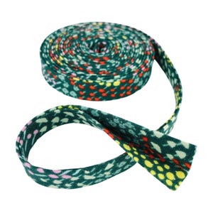 May include: A roll of dark green fabric bias tape featuring a repeating pattern of small, colorful dots in shades of red, yellow, and light pink. The tape is coiled into a circular shape with one end extending outward, showing the folded edges.