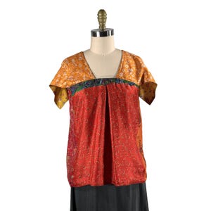 May include: A short-sleeved blouse featuring a V-neckline and a color-blocked design. The upper section is a golden yellow with a floral pattern, while the lower section is a vibrant red with a repeating intricate pattern, separated by a dark band.