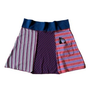 May include: A skirt featuring a wide navy blue elastic waistband and three distinct panels. The left panel has vertical stripes in red, white, and black. The center panel has navy blue diagonal stripes. The right panel has horizontal red stripes.