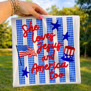 May include: White square pillow with blue and white striped and gingham pattern. The pillow has the text "She Loves Jesus and America too" in red script. Features an American flag, blue stars, and a Uncle Sam hat.