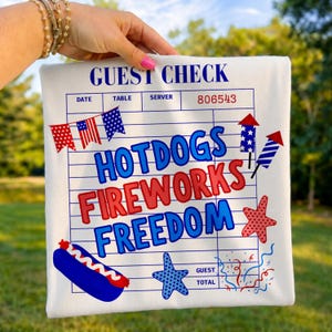 May include: White t-shirt with a graphic design resembling a guest check. The design includes the words "HOTDOGS FIREWORKS FREEDOM" in red and blue, along with illustrations of fireworks, a hotdog, and stars.