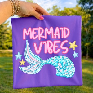 Może przedstawiać: Kwadratowy fioletowy przedmiot z tkaniny z napisem MERMAID VIBES dużymi r&oacute;żowymi literami. Pod napisem znajduje się grafika syreniego ogona z niebiesko-r&oacute;żowymi łuskami. Wz&oacute;r zawiera małe ż&oacute;łte i jasnoniebieskie gwiazdki rozrzucone wok&oacute;ł ogona.