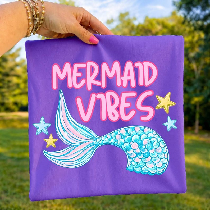 Może przedstawiać: Kwadratowy fioletowy przedmiot z tkaniny z napisem MERMAID VIBES dużymi r&oacute;żowymi literami. Pod napisem znajduje się grafika syreniego ogona z niebiesko-r&oacute;żowymi łuskami. Wz&oacute;r zawiera małe ż&oacute;łte i jasnoniebieskie gwiazdki rozrzucone wok&oacute;ł ogona.