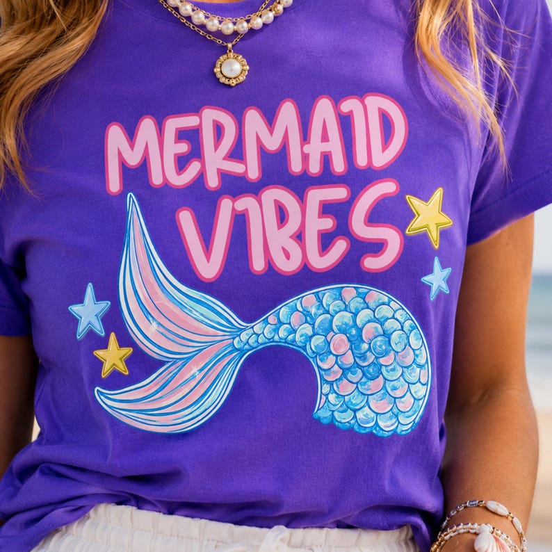 Może przedstawiać: Fioletowy t-shirt z napisem MERMAID VIBES r&oacute;żowymi literami. Pod napisem znajduje się grafika syreniego ogona z niebiesko-r&oacute;żowymi łuskami i płetwami. Wz&oacute;r zawiera małe ż&oacute;łte i jasnoniebieskie gwiazdki otaczające ogon.