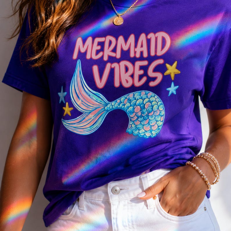 Może przedstawiać: Fioletowy t-shirt z napisem MERMAID VIBES r&oacute;żowymi literami nad grafiką przedstawiającą niebiesko-r&oacute;żowy ogon syreny. Wz&oacute;r zawiera małe ż&oacute;łte i niebieskie gwiazdki. Koszulka jest zestawiona z białymi szortami dżinsowymi ze srebrnym guzikiem.