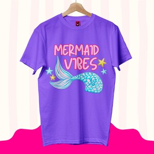 Może przedstawiać: Fioletowy t-shirt z kr&oacute;tkim rękawem z napisem "MERMAID VIBES" r&oacute;żowymi literami. Pod tekstem znajduje się grafika niebiesko-turkusowego ogona syreny z łuskami, otoczonego małymi ż&oacute;łtymi i jasnoniebieskimi gwiazdkami na przodzie koszulki.