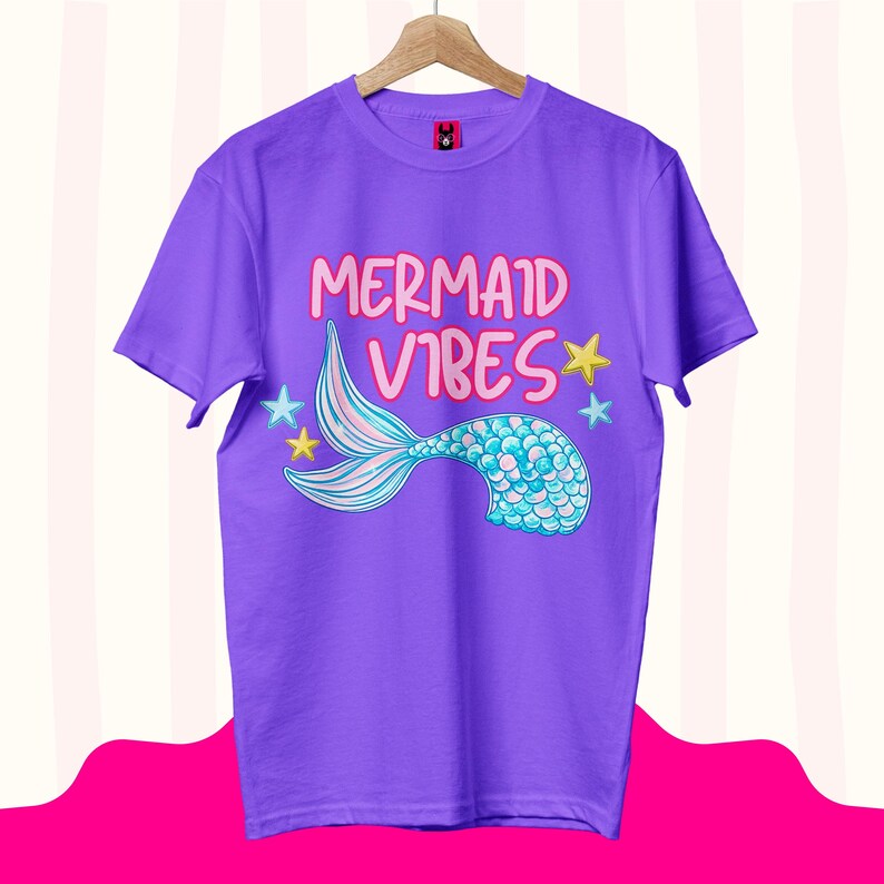 Może przedstawiać: Fioletowy t-shirt z kr&oacute;tkim rękawem z napisem "MERMAID VIBES" r&oacute;żowymi literami. Pod tekstem znajduje się grafika niebiesko-turkusowego ogona syreny z łuskami, otoczonego małymi ż&oacute;łtymi i jasnoniebieskimi gwiazdkami na przodzie koszulki.