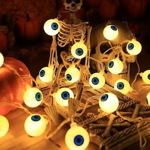 May include: A string of glowing LED lights shaped like white eyeballs with blue irises and black pupils. The lights are draped over a plastic skeleton and a skull, creating a bright, illuminated display for a festive seasonal decoration.