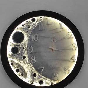 May include: A circular wall clock with a black frame and a textured, cratered surface on the left side resembling the moon. The clock face features a marbled gray finish with silver numerals from 1 to 12 and thin clock hands, illuminated by a soft light.