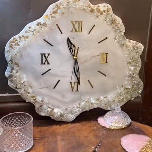 Può includere: Un orologio da parete decorativo con quadrante bianco e numeri romani dorati. Il bordo irregolare dell'orologio è adornato da dettagli dorati. Le lancette dell'orologio sono nere e dorate. L'orologio è su un tavolo di legno.