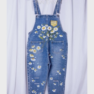 Può includere: Salopette in denim blu con motivo floreale a margherite bianche. La salopette ha spalline regolabili, una tasca frontale e dettagli strappati. Il testo "RXN JEANS" è visibile sull'appendiabiti.