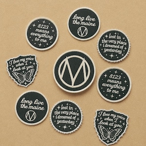 May include: A collection of black and white embroidered patches with various phrases and designs. The patches include text like "8123 means everything to me" and "long live the maine". Some patches feature a butterfly.